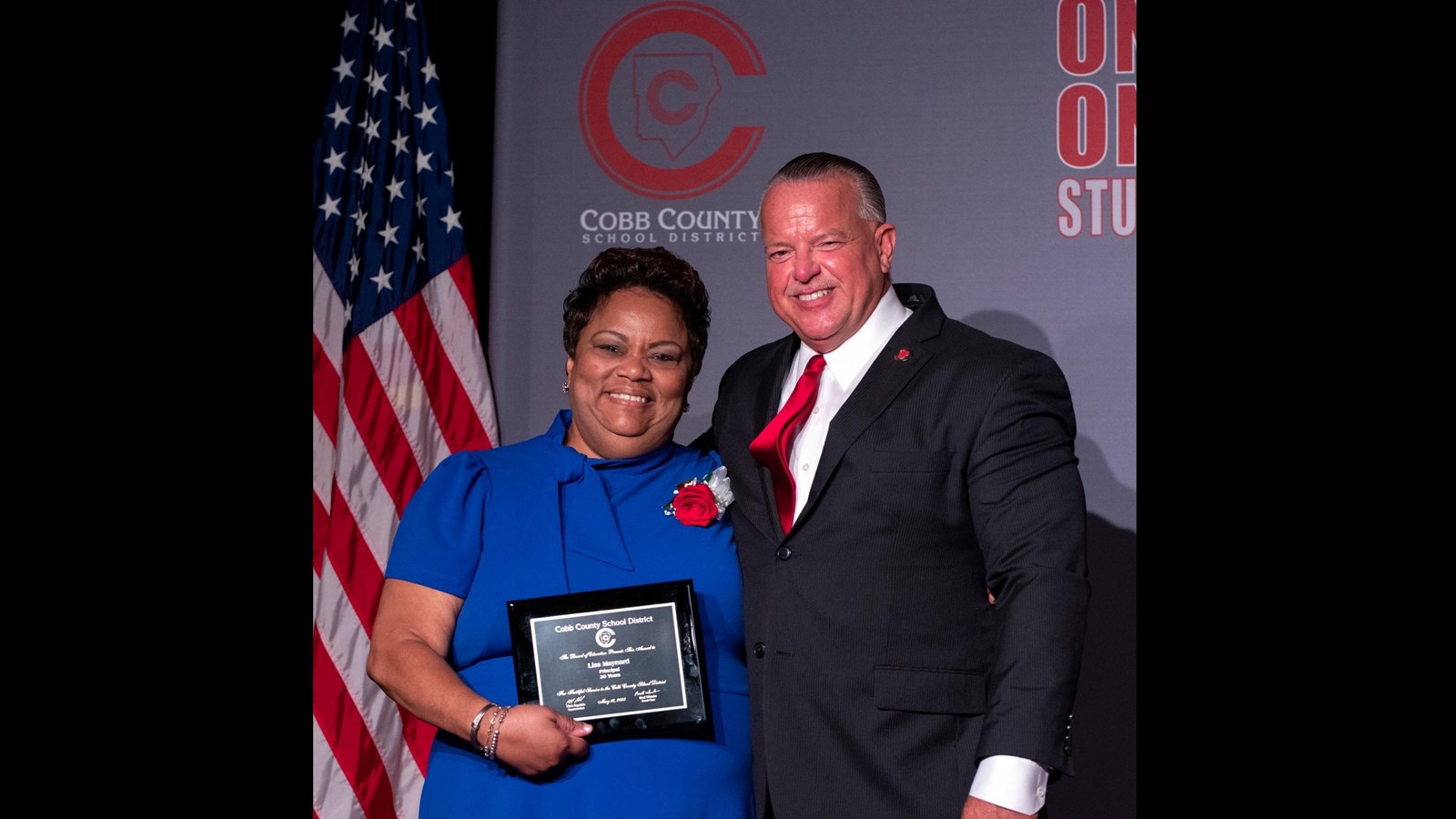 Liss Maynard in blue dress with Superintendent Ragsdale in front of an American flag and Cobb County logo
